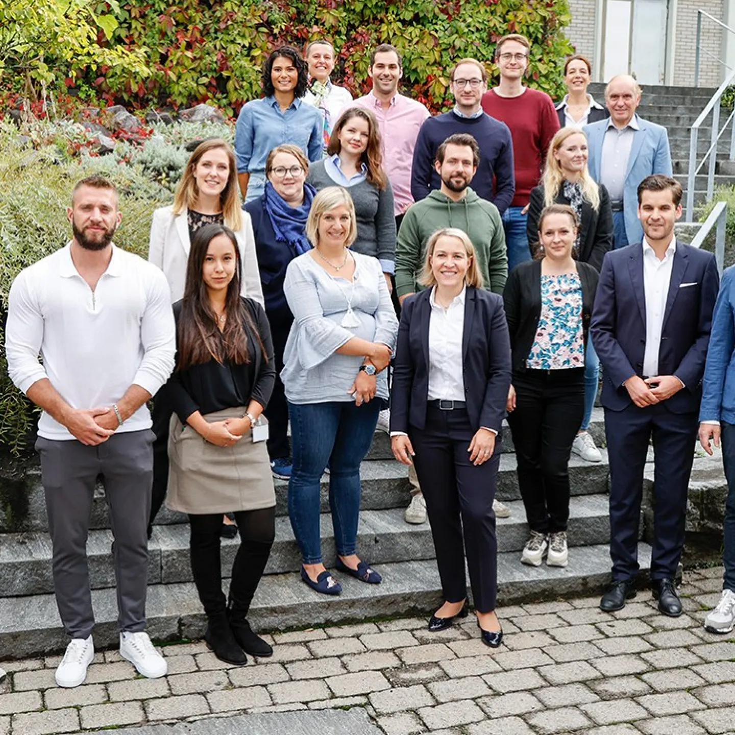 Eine Gruppe von Studierenden und Dozenten des Executive Master of Laws (LL.M.) im Gesellschafts-, Stiftungs- und Trustrecht steht auf einer Treppe im Freien. Sie lächeln in die Kamera, umgeben von grüner Vegetation und einem Gebäude im Hintergrund.
