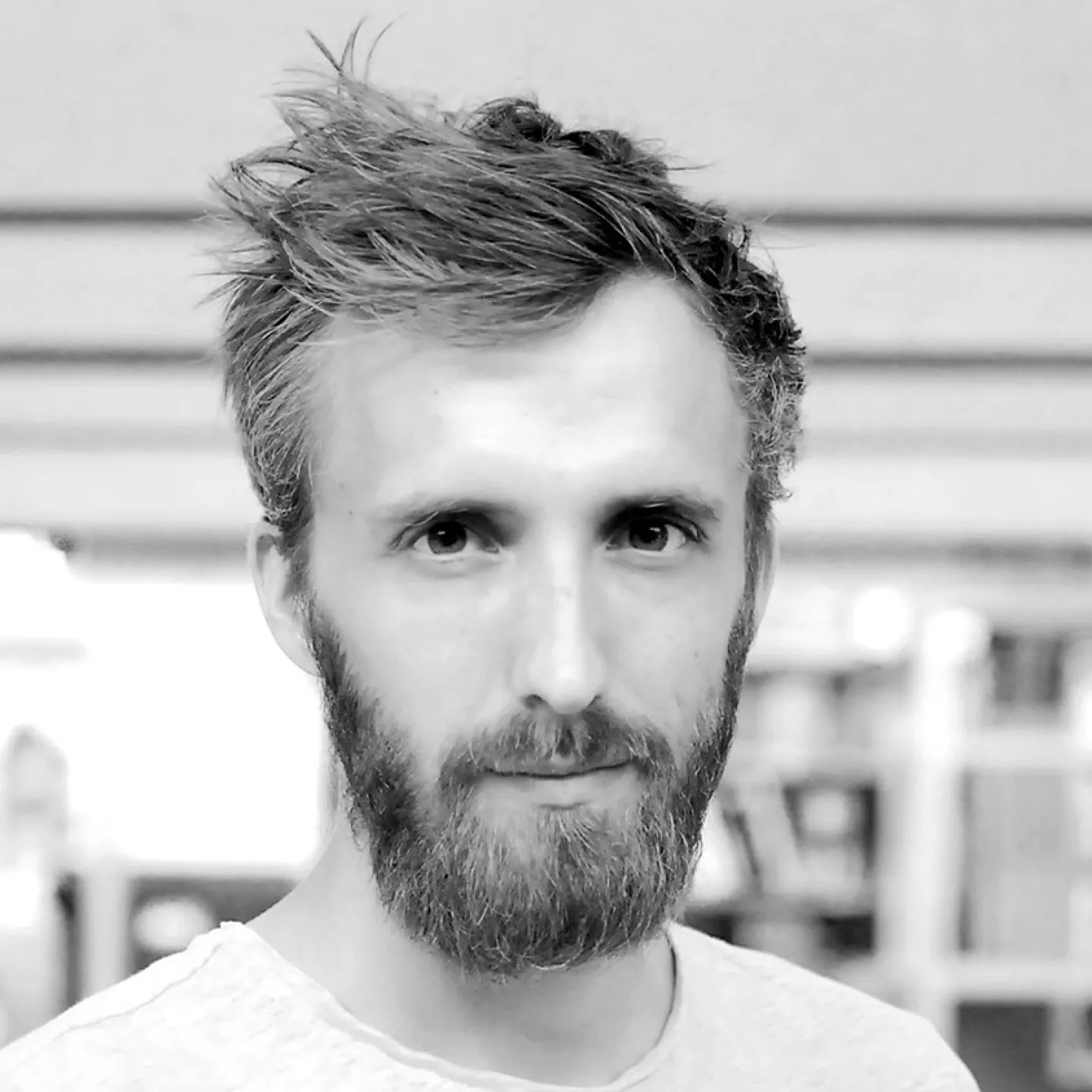 Ein Mann mit Bart und kurzem Haar blickt direkt in die Kamera. Der Hintergrund ist unscharf und zeigt eine Innenraumszene.