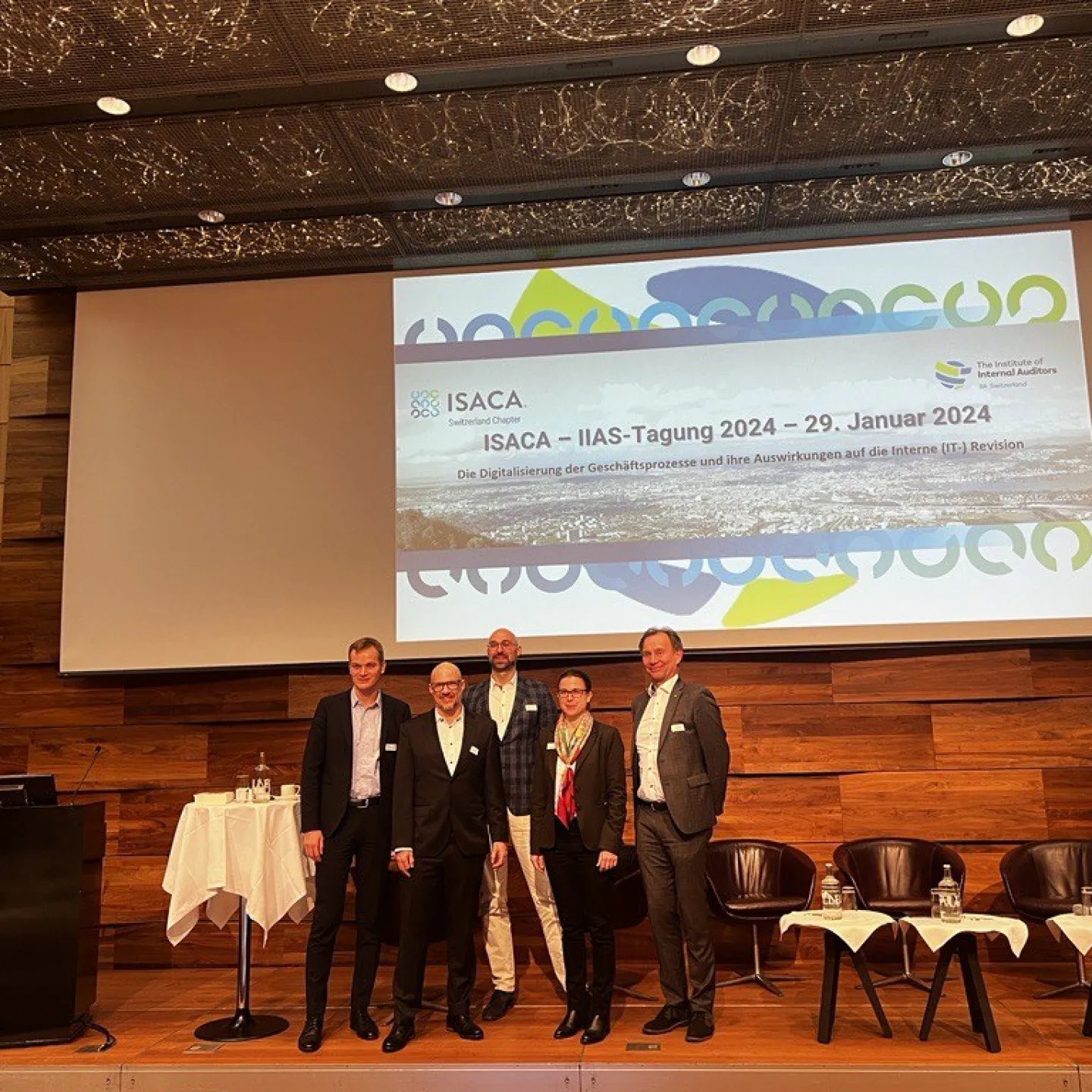 Gruppe von fünf Personen steht auf einer Bühne vor einer Leinwand mit dem Titel der ISACA-IIAS-Tagung 2024 in Zürich. Im Hintergrund sind Stühle und ein Tisch mit Wasserflaschen zu sehen.