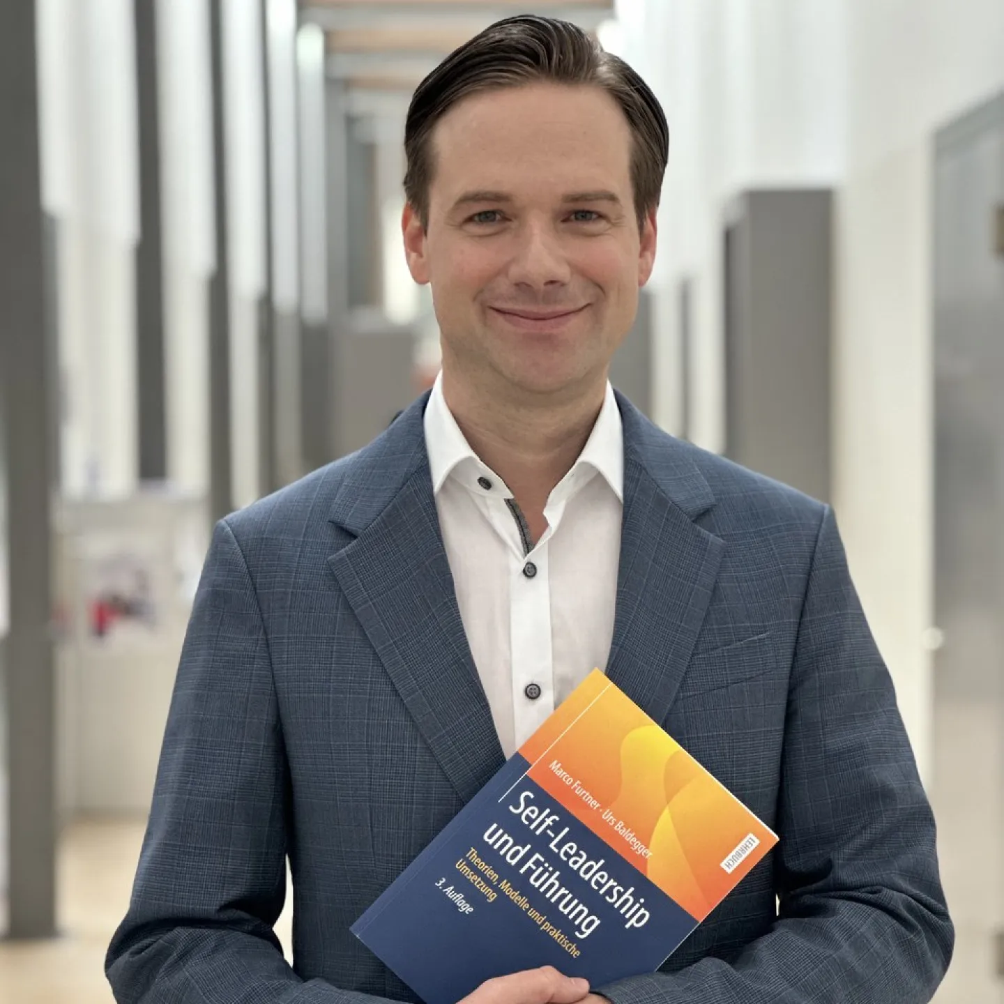 Ein Mann in einem blauen Anzug hält das Buch „Self-Leadership und Führung“ in einem modernen, hellen Flur.