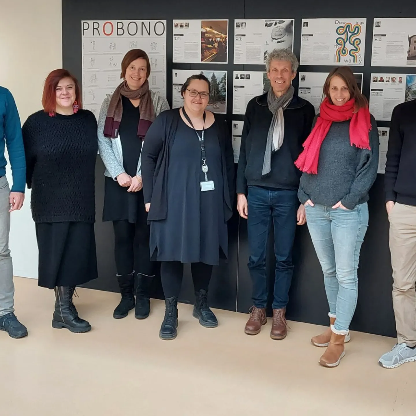 Gruppe von sieben Personen steht vor einer Wand mit dem Titel "PROBONO" und mehreren Plakaten. Sie lächeln in die Kamera und repräsentieren das Service Learning Netzwerk an der Universität Liechtenstein.