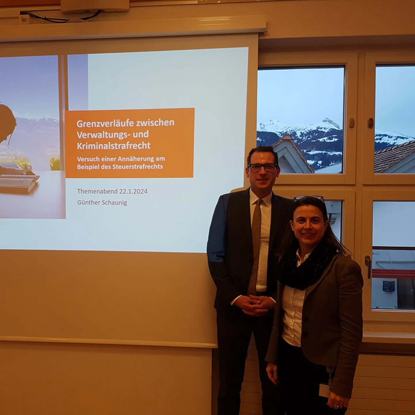 Ein Mann und eine Frau stehen vor einer Präsentationsleinwand mit dem Titel "Grenzverläufe zwischen Verwaltungs- und Kriminalstrafrecht" im Seminarraum der Universität Liechtenstein. Im Hintergrund sind schneebedeckte Berge durch die Fenster sichtbar.