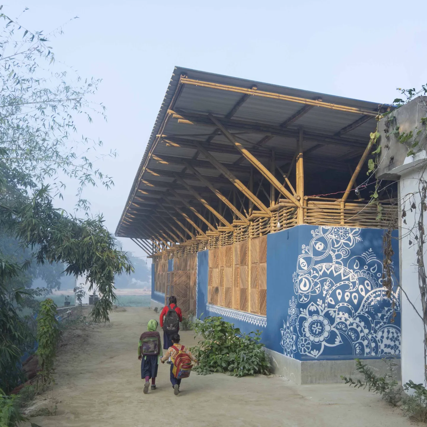 Kinder gehen auf einem sandigen Weg an einem blau bemalten Bambusgebäude mit weißem Ornamentmuster vorbei.