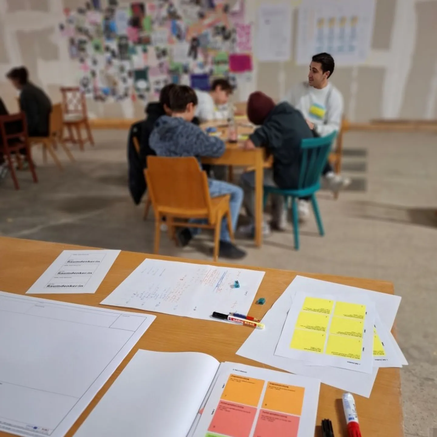 Blick auf einen Schreibtisch mit bunten Notizzetteln und Menschen, die an einem Tisch sitzen im Hintergrund