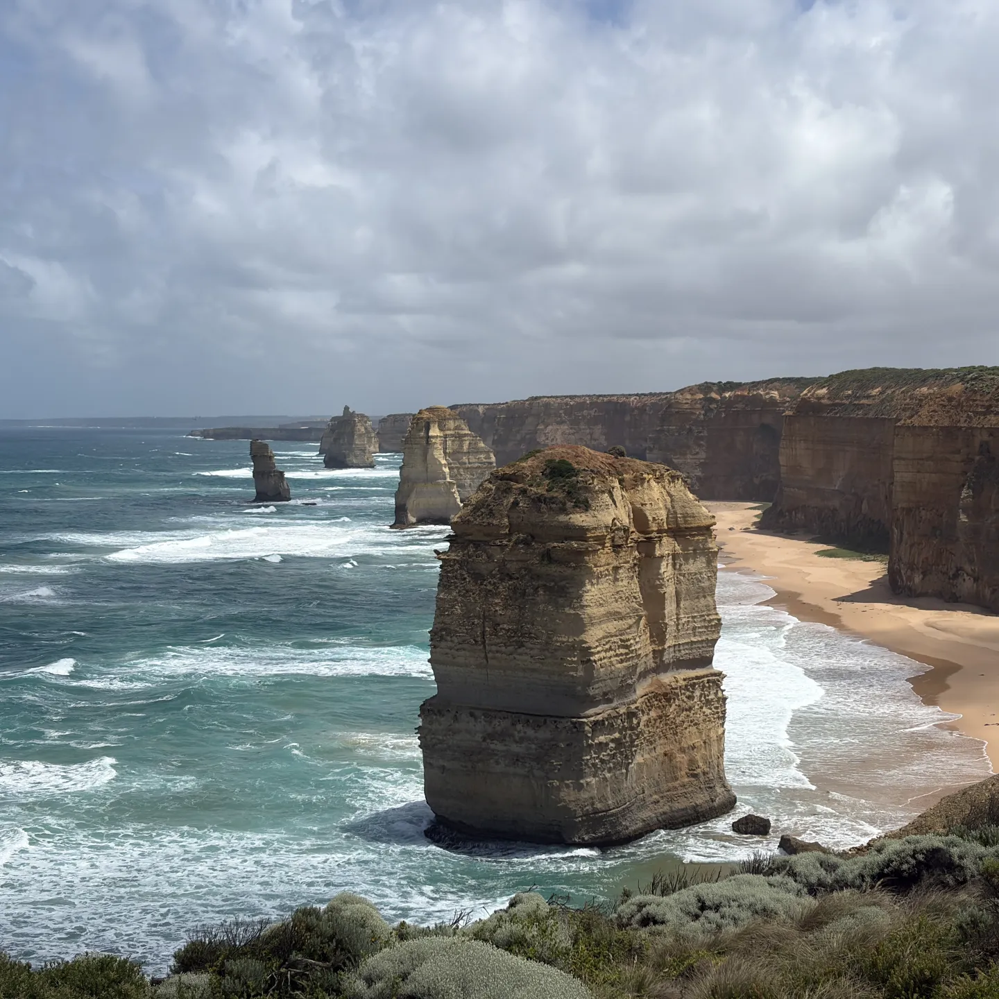 Steile Felsklippen und markante Felsnadeln ragen aus dem Meer entlang einer wilden Küste mit Sandstrand und brechenden Wellen.