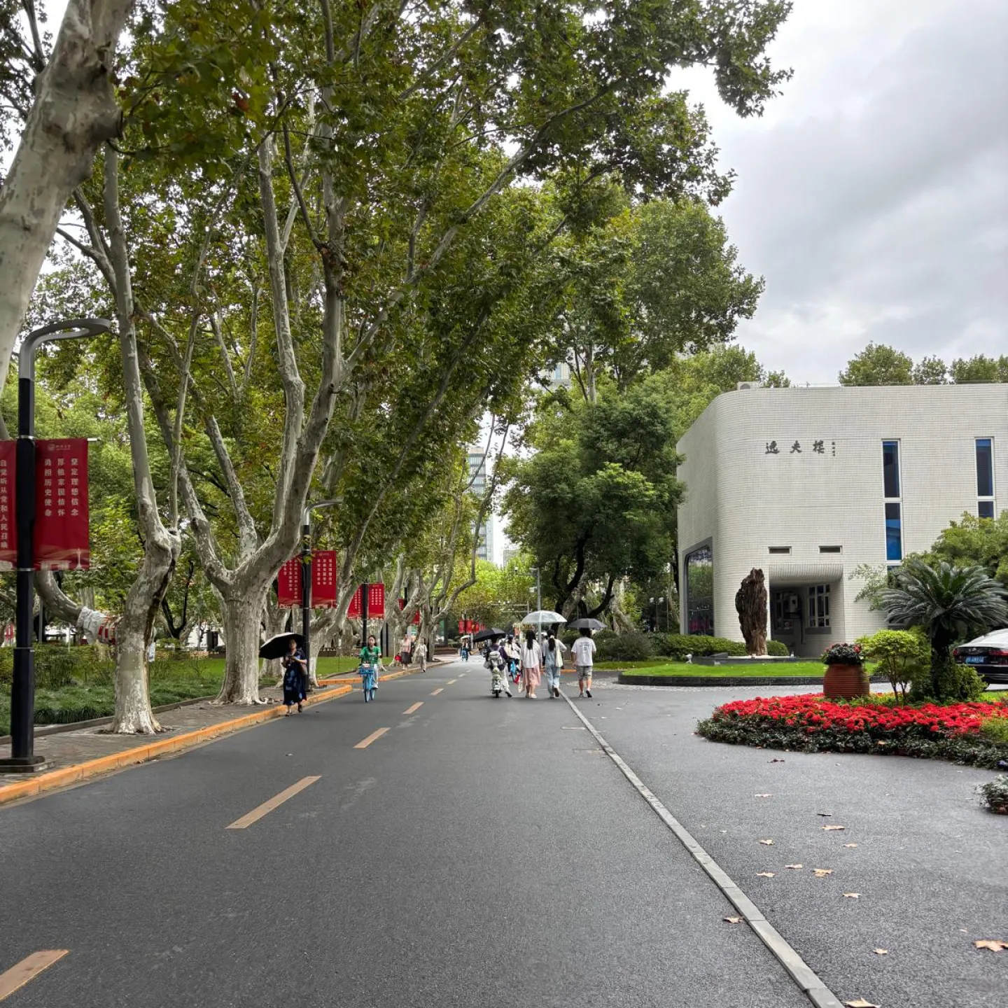 Baumgesäumte Allee auf einem Campus mit Spaziergängern, roten Bannern und einem modernen Gebäude am Straßenrand.