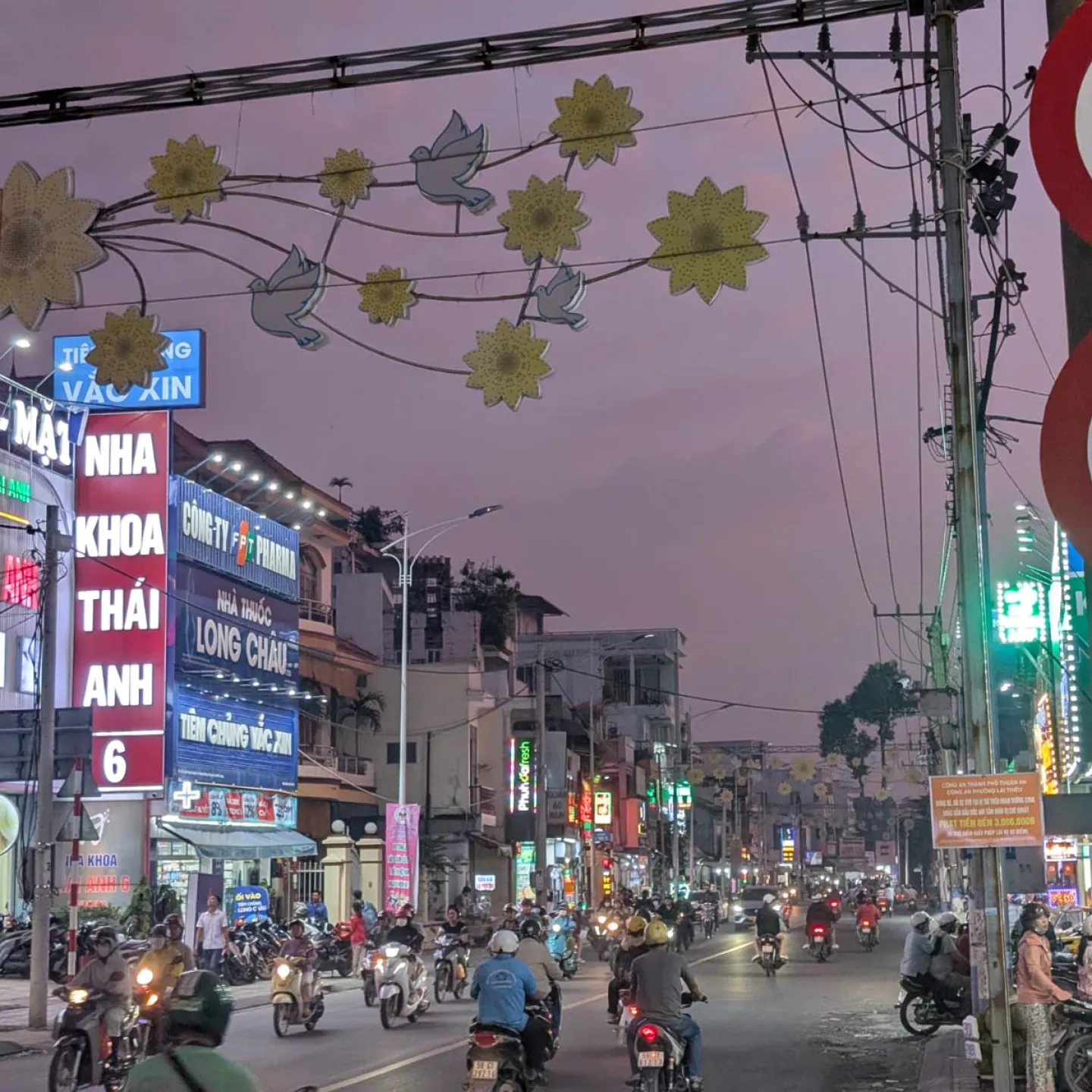 Belebte Straße bei Dämmerung mit vielen Motorrollern, leuchtenden Werbeschildern und dekorativen Lichtern über der Fahrbahn.