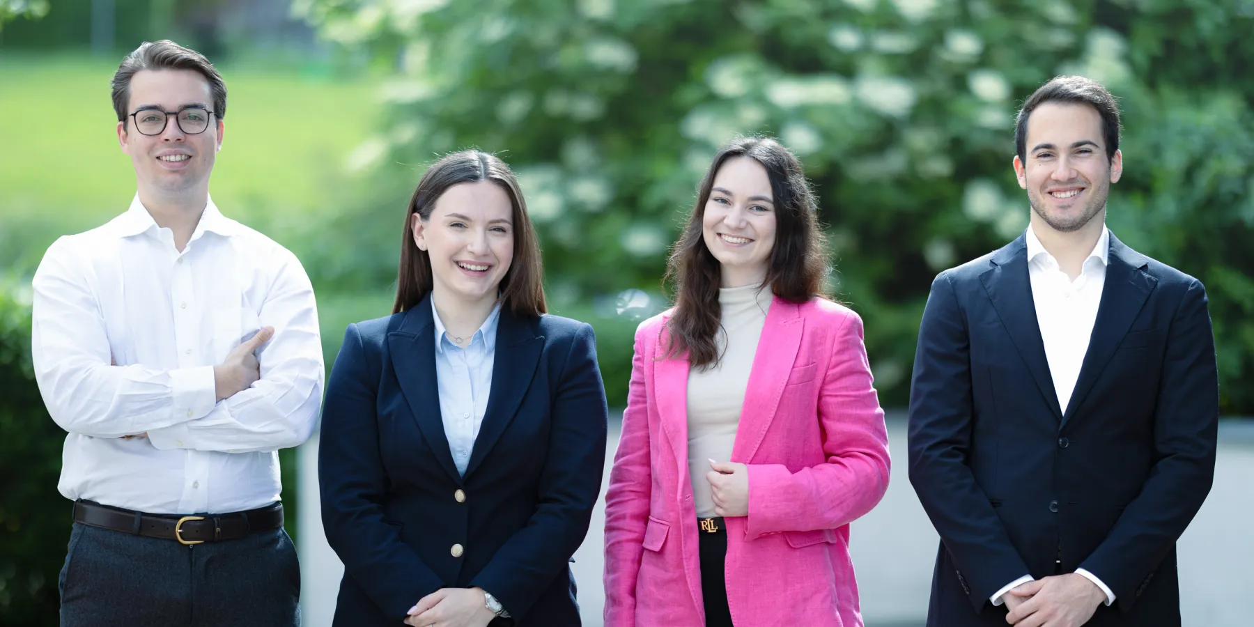 Vier Studierende im Masterstudiengang Innovative Finance der Universität Liechtenstein stehen nebeneinander im Freien, professionell gekleidet und lächelnd in die Kamera. Im Hintergrund ist grüne Natur zu sehen.