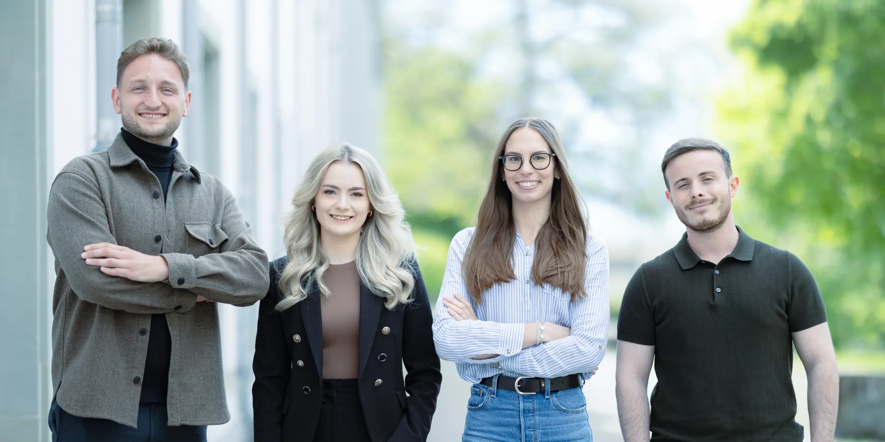 Vier Studierende im Masterstudiengang Information Systems der Universität Liechtenstein stehen nebeneinander auf einem Gehweg vor einem Universitätsgebäude und lächeln in die Kamera. Die Kleidung reicht von Business- bis Casual-Stil.