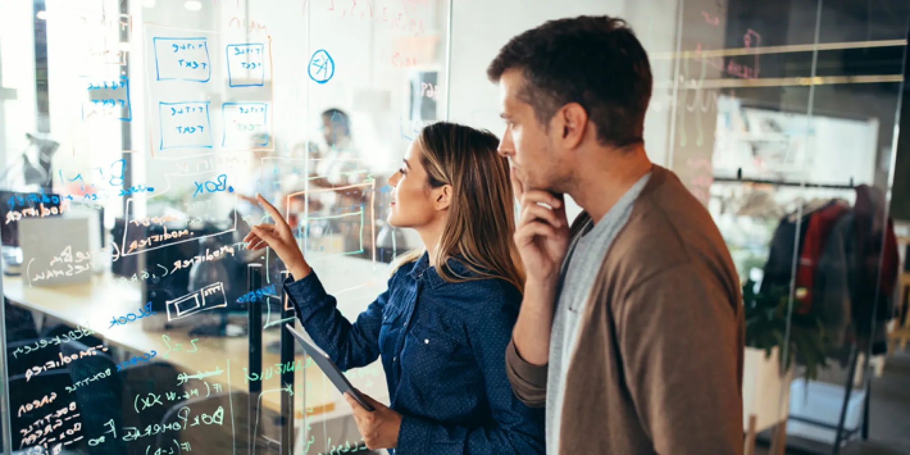 Ein Mann und eine Frau betrachten gemeinsam eine große Glaswand, die mit Notizen und Diagrammen beschrieben ist. Beide wirken nachdenklich und konzentriert, während sie an einer komplexen Aufgabe arbeiten.
