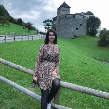 Studentin der Universität Liechtenstein sitzt vor dem Schloss Vaduz auf einem Holzzaun mit Blick in die Kamera, umgeben von grüner Hügellandschaft.