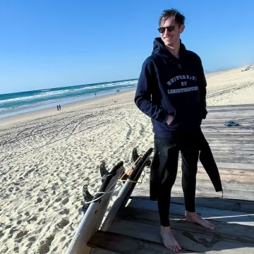 Student der Universität Liechtenstein steht barfuß am Strand mit Surfausrüstung und trägt einen Hoodie mit Universitätslogo während des Auslandssemesters.
