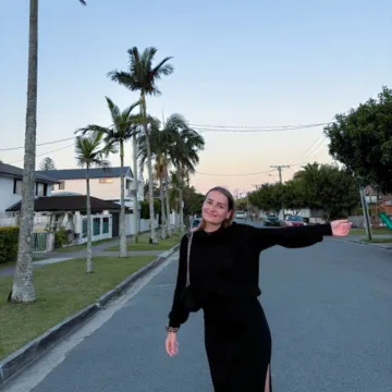 Studentin der Universität Liechtenstein steht lachend auf einer Straße mit Palmen in Australien und zeigt auf ihre Umgebung beim Auslandssemester.