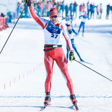 Ein Langläufer der Universität Liechtenstein überquert im roten Wettkampfanzug die Ziellinie eines Langlaufrennens im Schnee, jubelnd mit erhobenem Arm.
