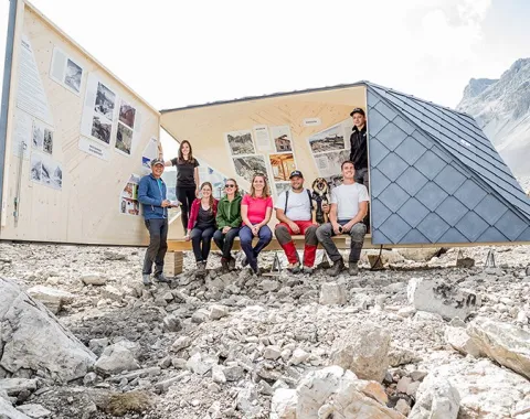 Eine Gruppe von Studierenden sitzt vor einer modernen, dreieckigen Bergunterkunft, die als architektonisches Ausstellungsprojekt dient und nachhaltige Baupraktiken in einer felsigen Berglandschaft zeigt.