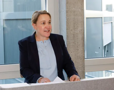 Eine Person hält eine Rede in einem modernen Gebäude. Sie steht hinter einem Rednerpult vor einem großen Fenster.