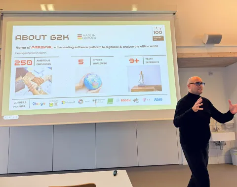 Ein Mann hält einen Vortrag vor einer Präsentationsfolie. Die Folie zeigt Informationen über das Unternehmen G2K, darunter die Anzahl der Mitarbeiter, Standorte und Jahre Erfahrung. Logos von Partnerunternehmen sind ebenfalls sichtbar.