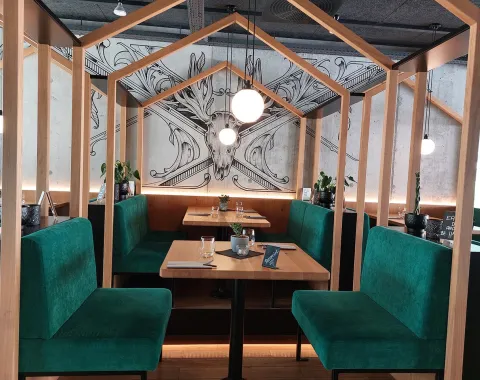 Ein stilvoll eingerichteter Restaurantbereich mit grünen Polsterbänken und Holztischen, umgeben von einem hölzernen Rahmen. Im Hintergrund ist eine kunstvolle Wandzeichnung eines Hirschkopfes zu sehen. Dezente Beleuchtung schafft eine einladende Atmosphäre.
