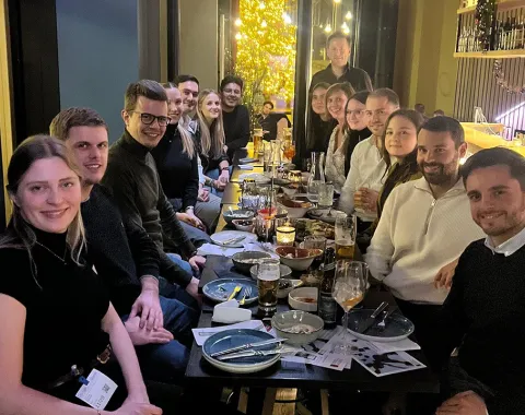 Eine Gruppe von Alumni der Universität Liechtenstein sitzt in einem Restaurant an einem langen Tisch, lächelnd und in geselliger Runde. Auf dem Tisch stehen Getränke und Teller mit Speisen. Die Atmosphäre ist warm und einladend.
