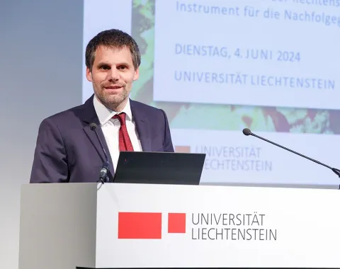 Ein Mann in einem Anzug steht an einem Rednerpult mit dem Logo der Universität Liechtenstein. Im Hintergrund ist eine Präsentationsfolie mit dem Datum 4. Juni 2024 zu sehen.