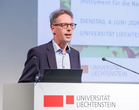 Ein Mann hält einen Vortrag im Auditorium der Universität Liechtenstein. Im Hintergrund ist eine Präsentation mit dem Datum 4. Juni 2024 zu sehen. Ein Rednerpult mit dem Logo der Universität steht vor ihm.
