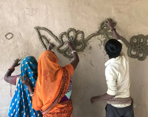 Drei Personen gestalten gemeinsam eine Wand mit traditionellen Mustern aus Lehm. Sie tragen bunte Kleidung und arbeiten konzentriert an einem floralen Design.