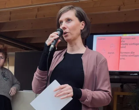 Eine Person hält ein Mikrofon und spricht vor einem Publikum. Sie trägt eine rosa Jacke und hält ein Blatt Papier. Im Hintergrund ist eine Präsentation auf einem Bildschirm zu sehen. Eine weitere Person steht links daneben und hört zu.