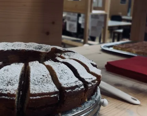 Ein frisch gebackener, mit Puderzucker bestäubter Kuchen steht auf einem Holztisch. Im Hintergrund sind Holzkonstruktionen und Tische in einem Raum zu sehen, die eine kreative und einladende Atmosphäre vermitteln.