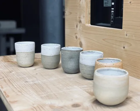 Sechs handgetöpferte Keramiktassen in verschiedenen Erdtönen stehen auf einem Holztisch. Die Tassen sind schlicht und modern gestaltet, passend zur Architekturveranstaltung an der Universität Liechtenstein.