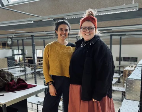 Zwei Personen stehen lächelnd in einem modernen Architekturatelier mit großen Fenstern und industriellem Design. Sie tragen lässige, kreative Kleidung. Im Hintergrund sind Arbeitsplätze und eine Uhr sichtbar.