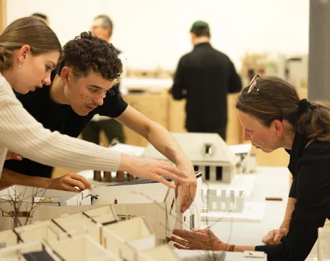Drei Personen betrachten und diskutieren über Architekturmodelle auf einem Tisch in einem Ausstellungsraum. Im Hintergrund sind weitere Personen und Modelle zu sehen.