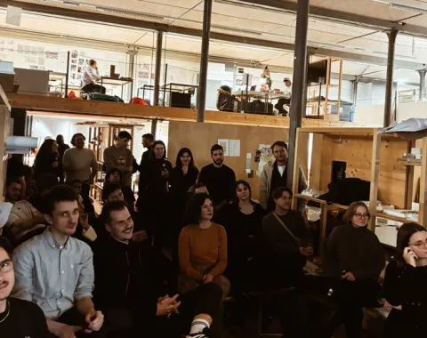 Eine Gruppe von Studierenden und Alumni sitzt in einem Architekturstudio und hört einer Präsentation zu. Im Hintergrund sind Arbeitsplätze und eine Uhr an der Wand zu sehen. Die Atmosphäre ist konzentriert und interessiert.