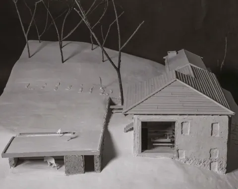 Ein Architekturmodell zeigt ein Gebäude mit Satteldach und angrenzendem offenen Unterstand auf einer schneebedeckten Landschaft. Im Hintergrund stehen stilisierte Bäume.