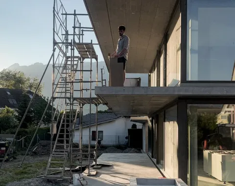 Ein Architekt steht auf einem modernen Gebäude mit großen Glasfenstern, daneben ein Baugerüst. Im Hintergrund sind Berge und ein Haus zu sehen. Die Szene zeigt eine Baustelle bei Sonnenuntergang.