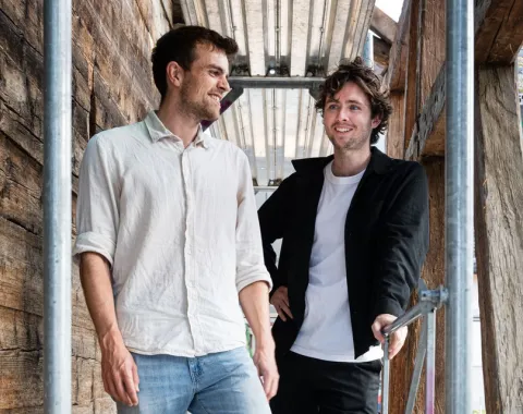 Zwei junge Männer stehen lächelnd auf einem Gerüst in einem Holzbau. Sie tragen legere Kleidung und wirken entspannt. Die Umgebung deutet auf eine Baustelle oder ein architektonisches Projekt hin.