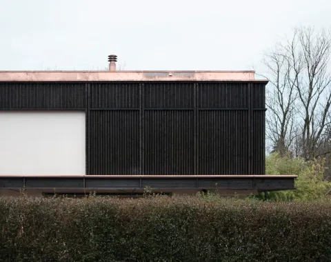 Ein modernes Gebäude mit dunkler Holzfassade und flachem Kupferdach, umgeben von Bäumen und einer Hecke im Vordergrund.