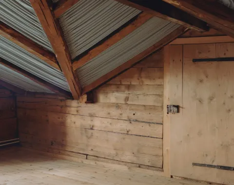 Innenansicht eines Dachbodens mit Holzwänden und Holzboden, sichtbare Dachbalken und Wellblechverkleidung. Eine Holztür befindet sich an der rechten Wand.