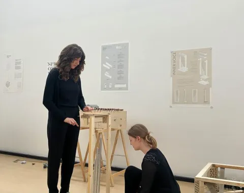 Zwei Studierende arbeiten in einem hellen Raum an einem architektonischen Modell. Im Hintergrund sind Poster mit Designplänen an der Wand zu sehen.