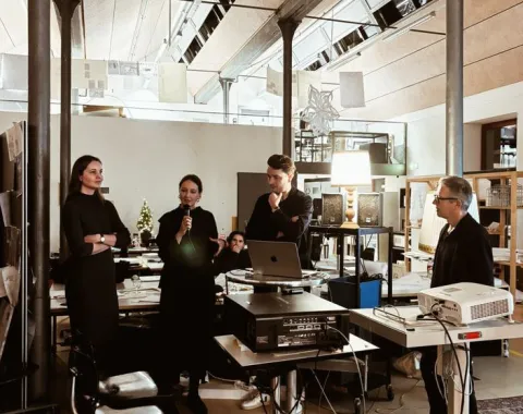Eine Gruppe von Personen steht in einem modernen Architekturstudio. Eine Person spricht in ein Mikrofon, während andere aufmerksam zuhören. Im Raum sind technische Geräte und Präsentationsmaterialien zu sehen.