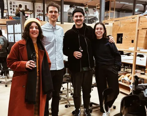 Vier Personen stehen lächelnd in einem Architekturstudio. Sie tragen lässige Kleidung und halten Getränke in der Hand. Im Hintergrund sind Arbeitsplätze und Modelle zu sehen. Die Atmosphäre wirkt freundlich und kreativ.