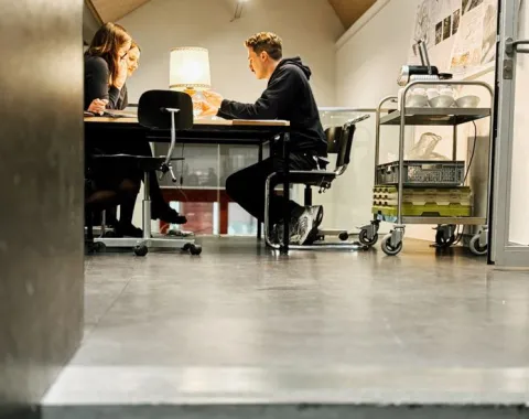 Drei Personen sitzen an einem Tisch in einem modernen Seminarraum und diskutieren. Im Hintergrund sind Architekturpläne an der Wand sichtbar. Der Raum ist hell beleuchtet und wirkt einladend.