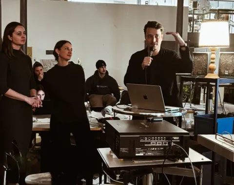 Eine Gruppe von Personen steht in einem Raum mit moderner Einrichtung. Eine Person hält ein Mikrofon und spricht, während andere zuhören. Im Hintergrund sind Tische, Stühle und technische Geräte zu sehen. Die Szene wirkt wie eine Präsentation oder ein Workshop in einem akademischen Umfeld.