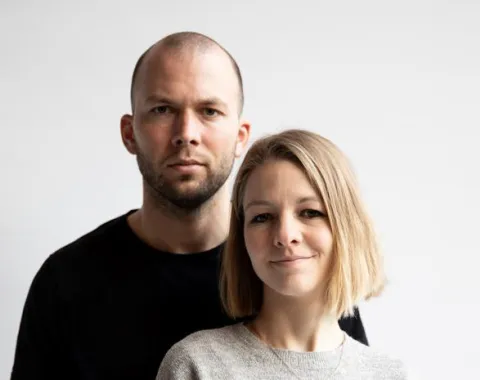 Ein Mann und eine Frau stehen vor einem neutralen Hintergrund. Beide blicken direkt in die Kamera. Die Frau trägt einen grauen Pullover, der Mann ein schwarzes T-Shirt.