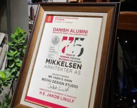 Ein gerahmtes Zertifikat zur Anerkennung der Zusammenarbeit im Bereich Architektur zwischen Dänemark und Pakistan, ausgestellt an Fasih D. Khan, steht auf einem Regal. Im Hintergrund sind Architekturprojekte und Gruppenfotos zu sehen.