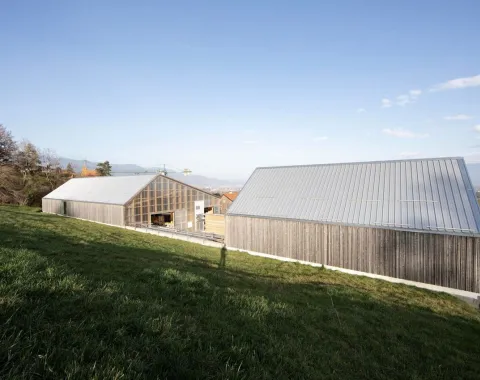 Zwei moderne Gebäude mit Holz- und Glasfassaden in einer ländlichen Umgebung. Die Architektur ist schlicht und kontextbezogen, eingebettet in eine grüne Landschaft.