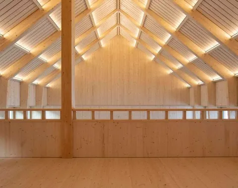 Innenansicht eines modernen Holzgebäudes mit sichtbarer Dachkonstruktion und natürlicher Beleuchtung. Die Architektur betont klare Linien und nachhaltige Materialien.