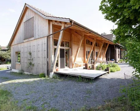 Ein modernes Holzgebäude mit schrägen Stützen und einer Terrasse, umgeben von Kies und Grünflächen, im ländlichen St. Galler Rheintal.
