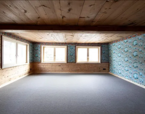 Ein leerer Raum mit Holzbalkendecke und Holzwänden, teilweise mit blauer, gemusterter Tapete. Drei große Fenster lassen Tageslicht herein. Der Boden ist mit grauem Teppich ausgelegt.
