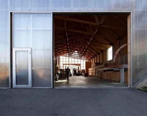 Ein modernes Gebäude mit einer großen offenen Tür, die den Blick auf eine helle, hölzerne Innenstruktur freigibt. Im Inneren sind Personen und gestapelte Holzmaterialien sichtbar. Die Architektur betont klare Linien und natürliche Materialien.