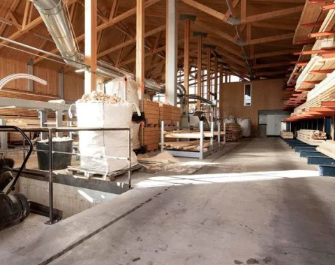Innenansicht einer modernen Holzwerkstatt mit sichtbaren Holzbalken und gestapelten Holzplanken. Die Werkstatt ist hell erleuchtet und zeigt eine aufgeräumte, funktionale Arbeitsumgebung.